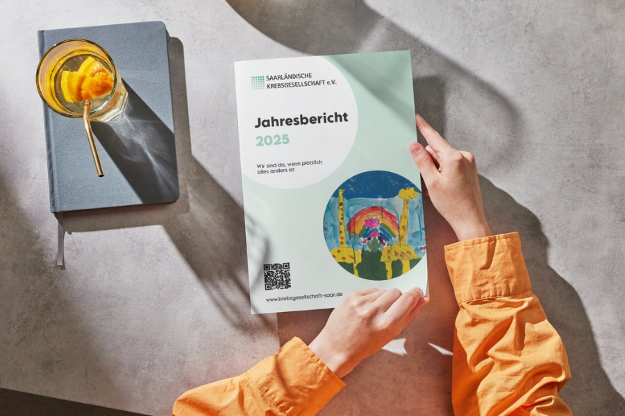 Eine Person mit einem orangefarbenen Hemd, von der man nur die Arme sieht, hält den Jahresbericht 2025 in den Händen